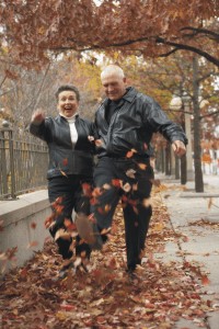 A couple of seniors, a man and a woman, kick and laugh their way through fallen leaves during an autumn walk.